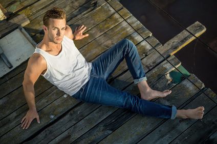 Cute barefoot guy lies on the wooden surface outdoors. He wears a white singlet and blue jeans and looks into the camera while leans on his hands. Photographed from the upper point. Horizontal.